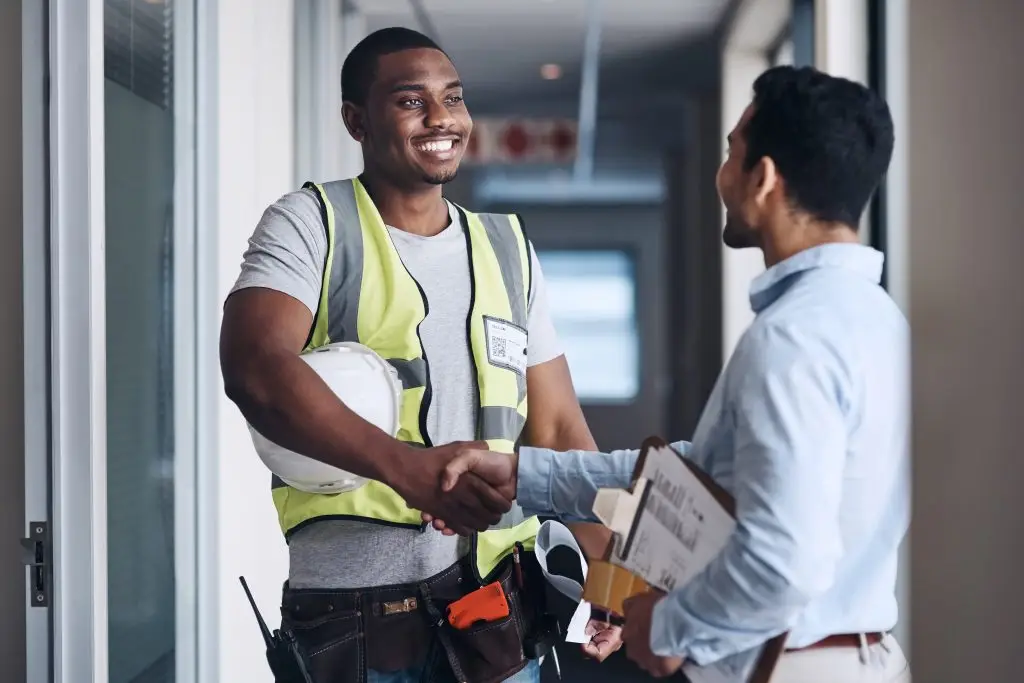 Smiling Black contractor in a high-visibility safety vest and hard hat holding blueprints while shaking hands with a client at a doorway.
