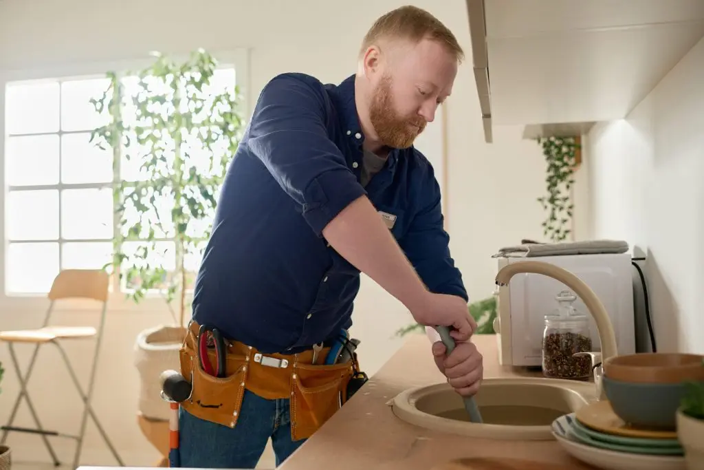 Bearded repairman wearing a blue shirt and tool belt using a plunger to clear a clogged kitchen sink.