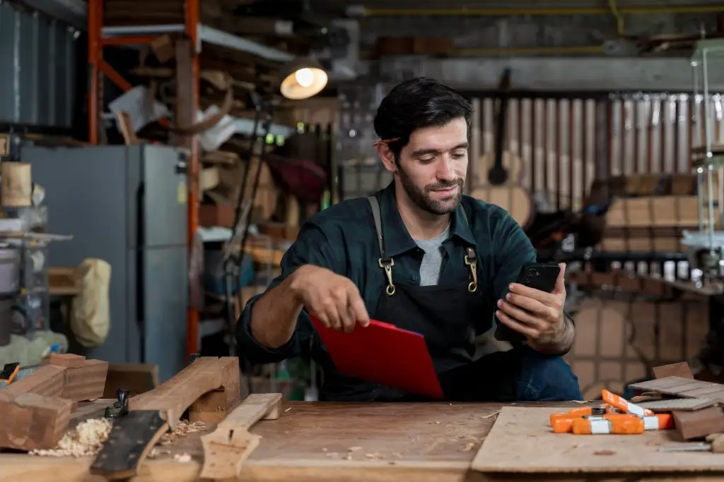 Male woodworker wearing a black apron with a pencil behind his ear using a smartphone at a workshop workbench next to a red clipboard.