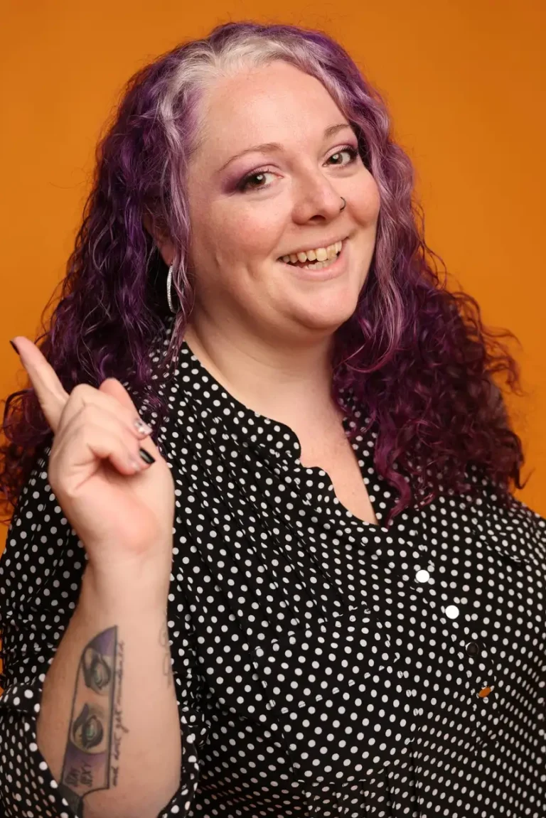 Celia Surridge, Founder & CEO of Capital Valley Marketing, smiling with vibrant purple hair, pointing upward while wearing a black-and-white polka dot blouse against an orange background.