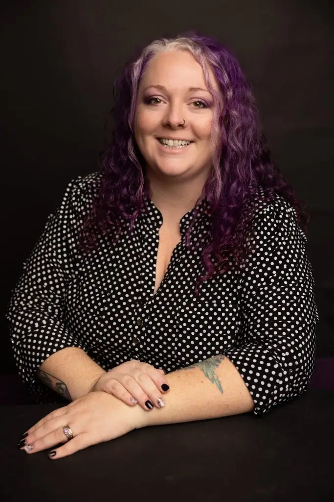 Celia Surridge, Founder & CEO of Capital Valley Marketing, smiling with arms crossed on a table in a patterned shirt with vibrant purple curly hair.