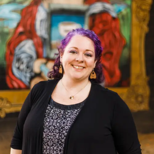 Headshot of Celia Surridge, Founder & CEO of Capital Valley Marketing, a smiling woman with vibrant purple hair wearing a black cardigan, pattern top, and gold earrings in front of framed artwork.