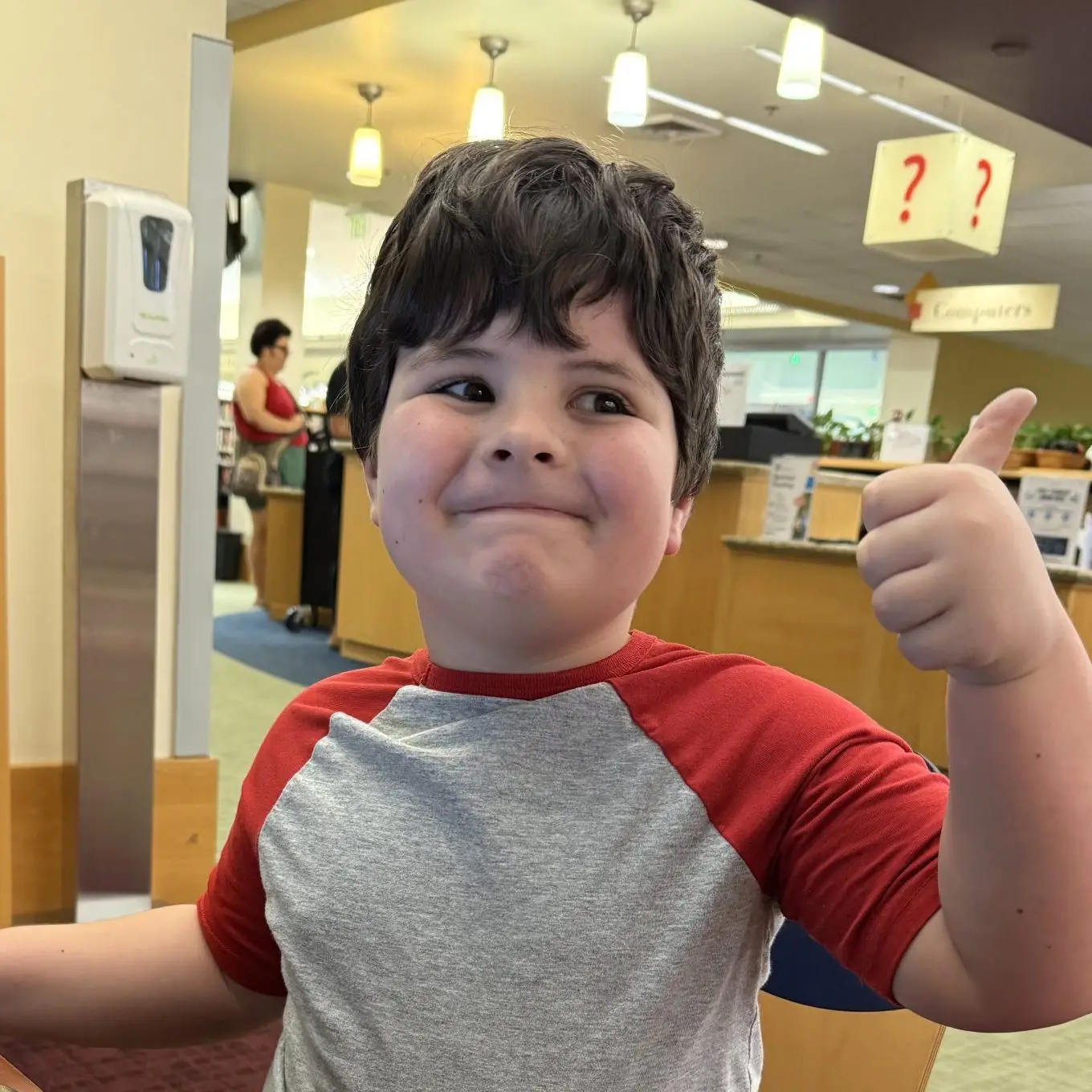 Young boy in a red and grey baseball shirt giving a thumbs-up gesture indoors.